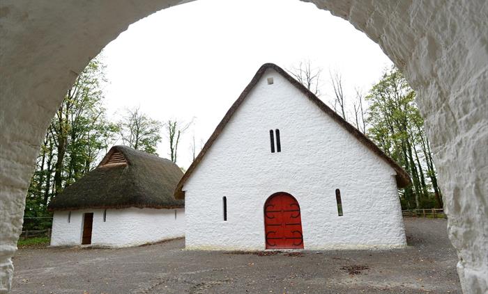 looking through a stone archway, you can see two white buildings with thatched rooves. The larger building has a big red door with black metalwork.